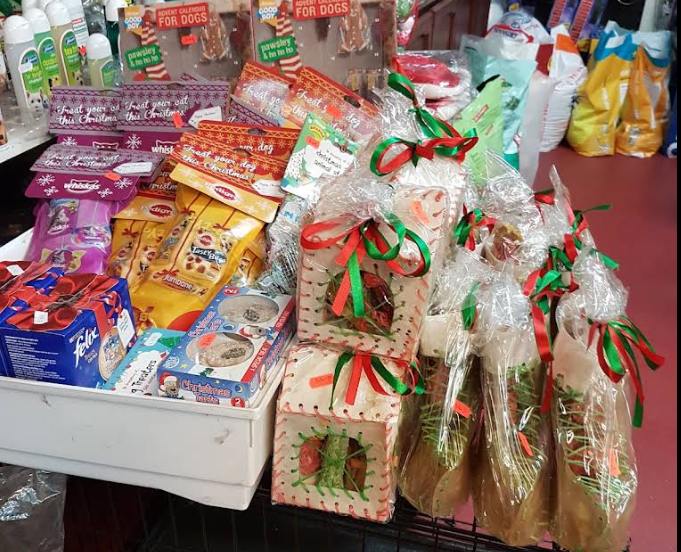 A retail display shelf filled with packaged dog treats and holiday-themed gift bags wrapped in clear cellophane with red and green ribbons. Visible packages include colorful pet food and treat boxes, with some labels partially legible such as 'K&H' and 'Purina'.